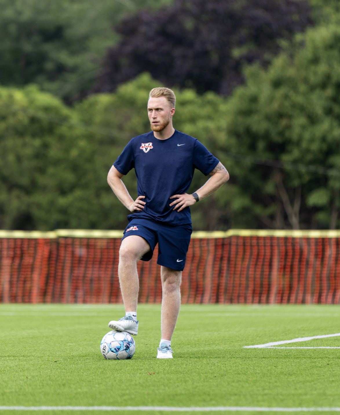 A middle-aged man with gray facial hair, wearing a brown polo shirt and cream-colored pants, sits on a bench in a room with neutral-colored walls.