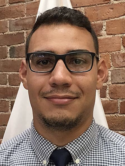 Close-up of a man with glasses, short dark hair, and a goatee, smiling in front of a brick wall.