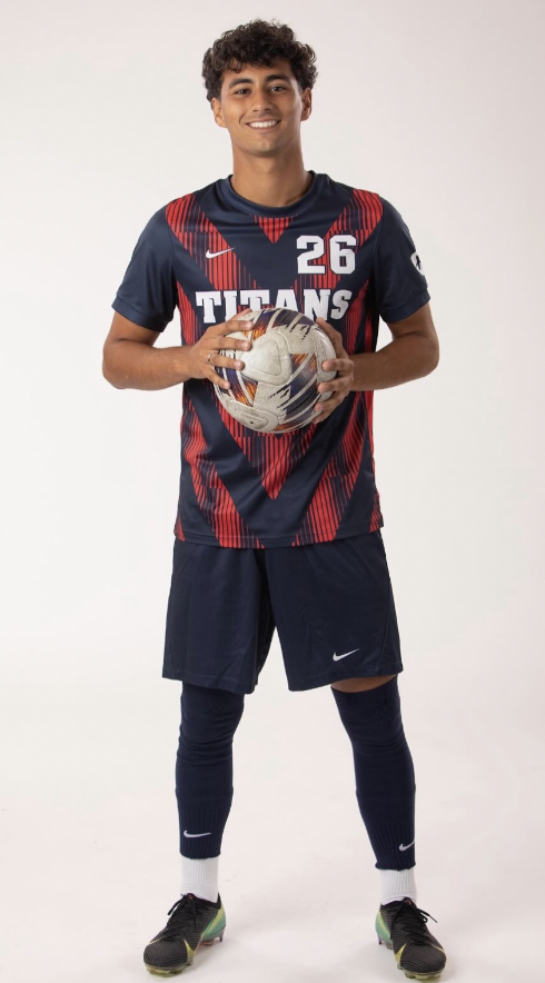 Young male soccer player holding a soccer ball, wearing a navy blue and red jersey with the number 26, shorts, knee-high compression socks, and athletic shoes, standing against a plain white background.