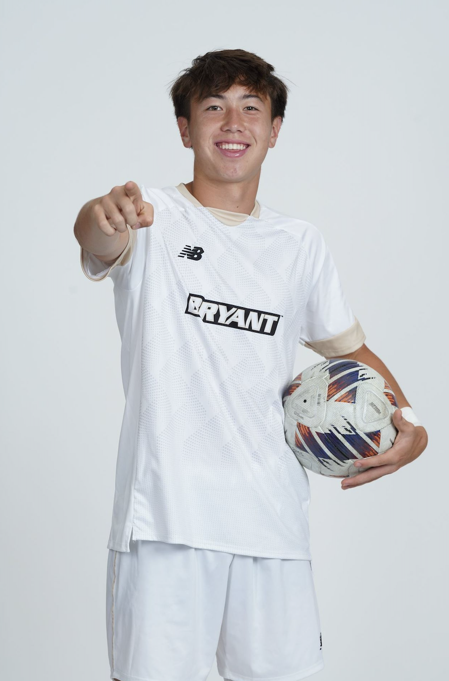A young male soccer player in a white jersey holding a soccer ball and pointing at the camera, smiling.