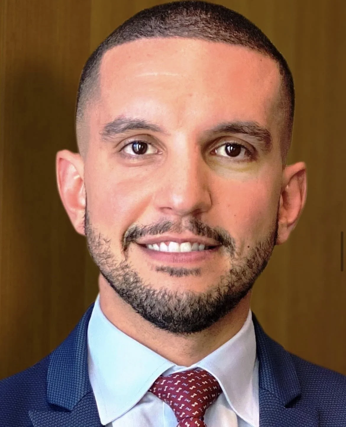 A man in a suit and tie smiling, with a short, well-groomed beard and dark hair