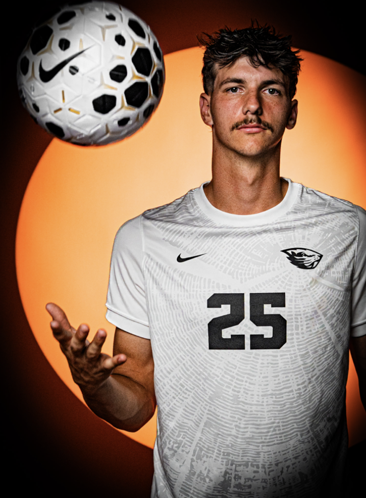 A young male soccer player with light skin and brown hair taking a selfie, holding a white and blue soccer ball on his shoulder, wearing a black and orange jersey with the number 25, in front of a sports-themed background.