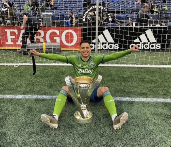 A soccer player in a green uniform sitting on the field with a large trophy between his legs, smiling, in front of a goal net with advertising banners in the background.