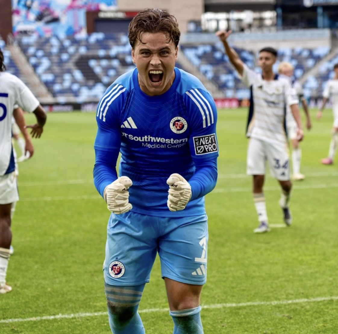 A soccer goalie in a blue uniform celebrating on a soccer field with clenched fists and an excited expression, with teammates in the background.