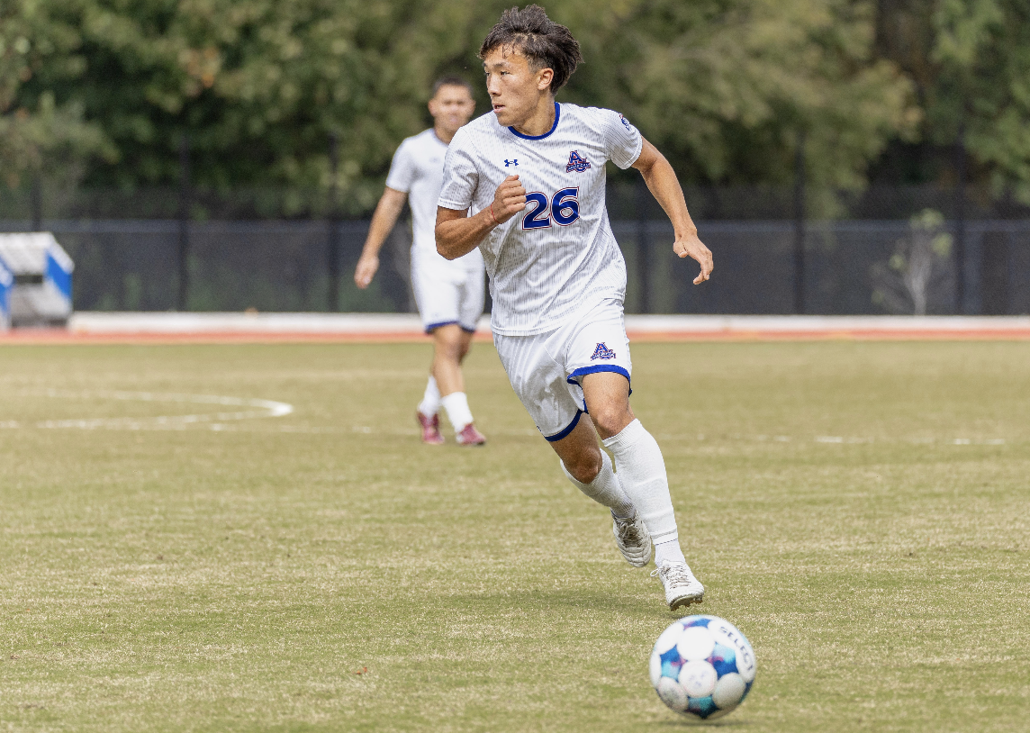 A soccer player in a white uniform with the number 26 dribbling a soccer ball on the field, with another player in the background.
