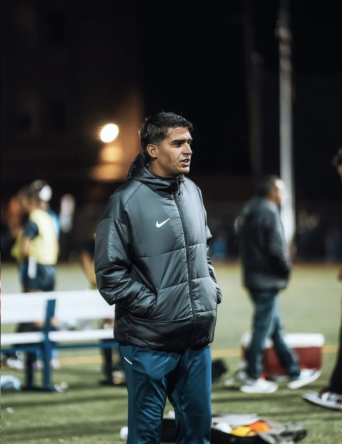 A man wearing a black Nike jacket with hands in pockets standing on a sports field at night with people and equipment in the background.