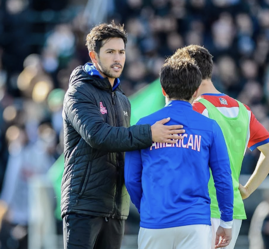 A man in a black jacket comforting a young boy in a blue jacket at a sports event, with other people in the background.