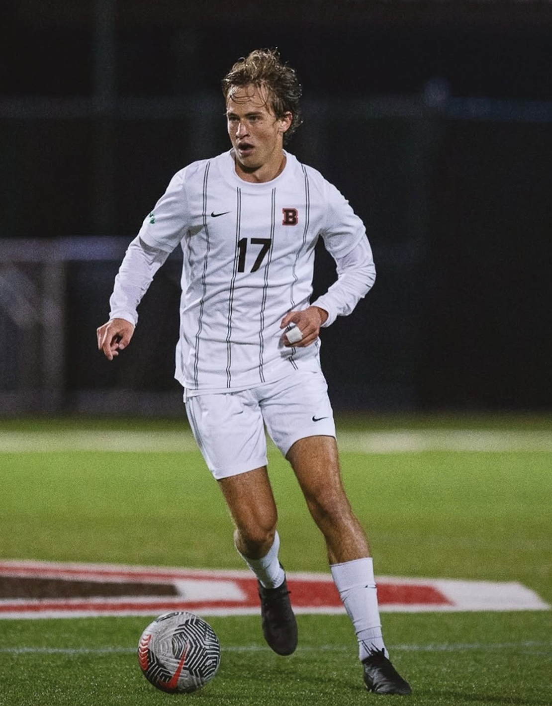 A male soccer player wearing a white uniform with black vertical stripes, number 17, is running on the field with a soccer ball at his feet during a night game.