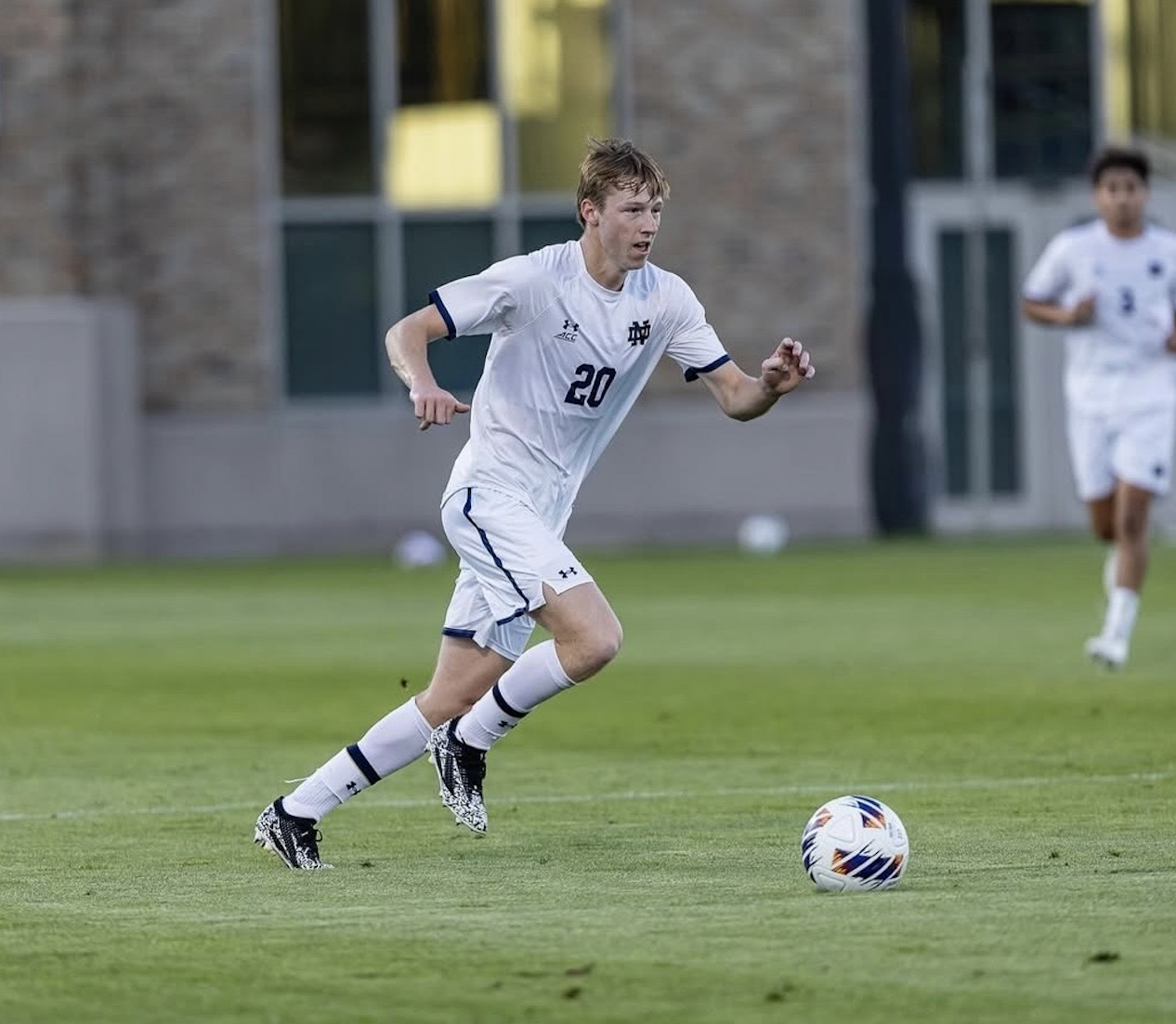 A young male soccer player in a white uniform with the number 20 running on a grass field with a soccer ball.