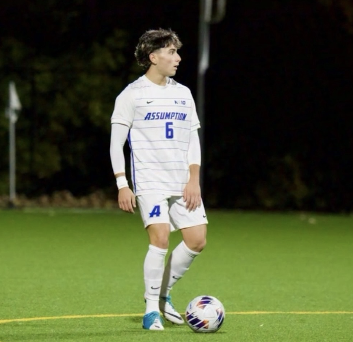 A soccer player in a white uniform with blue lettering and the number 6 stands on a green field preparing to kick a soccer ball at night.