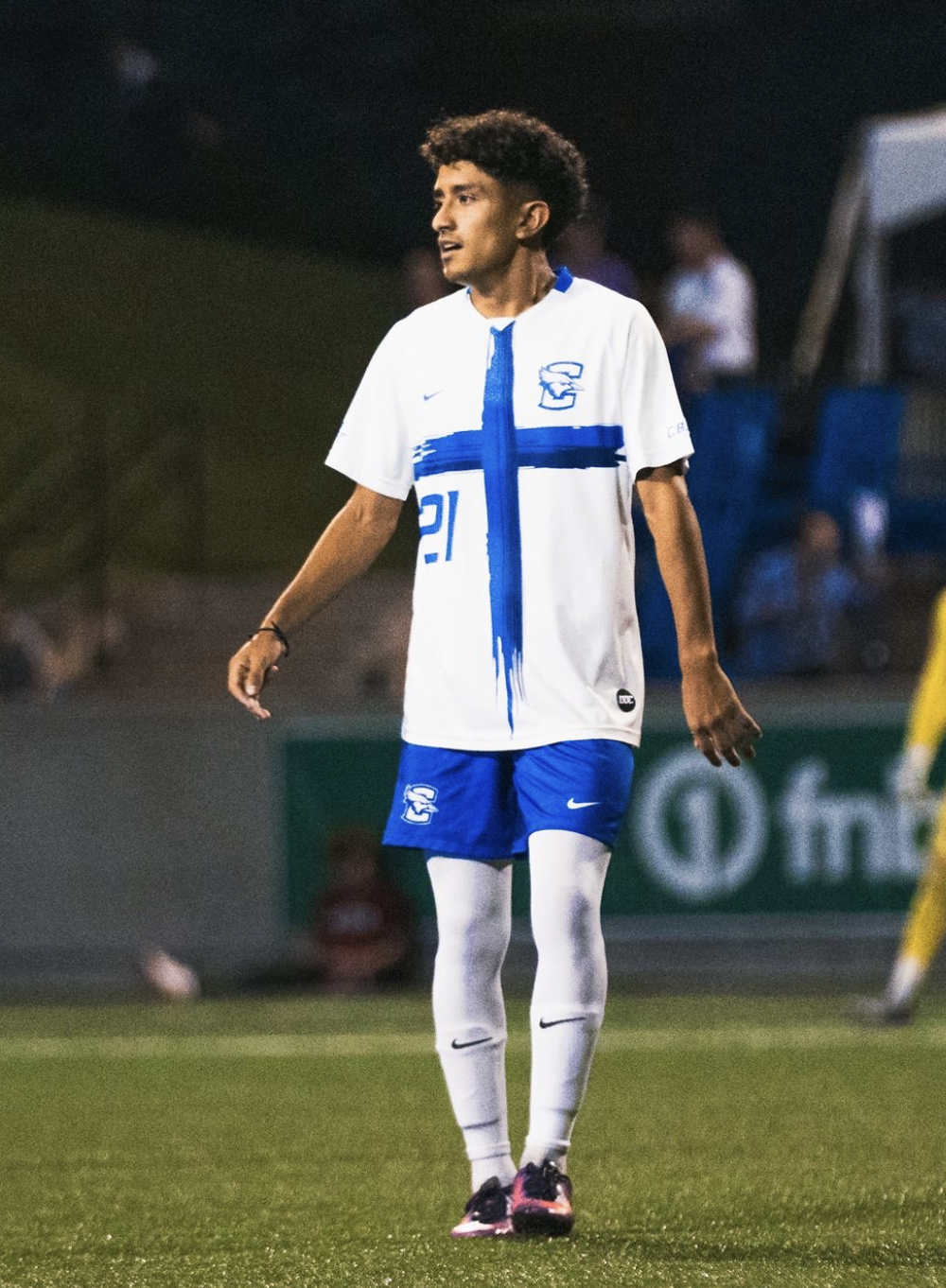 A young male soccer player with curly hair wearing a white and blue uniform, standing on a soccer field during a game.