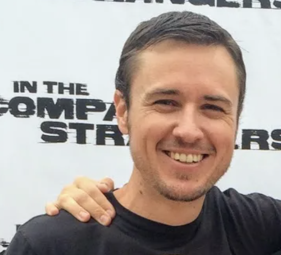 A smiling man with short brown hair and a light beard, wearing a black shirt, standing in front of a white background with black text.