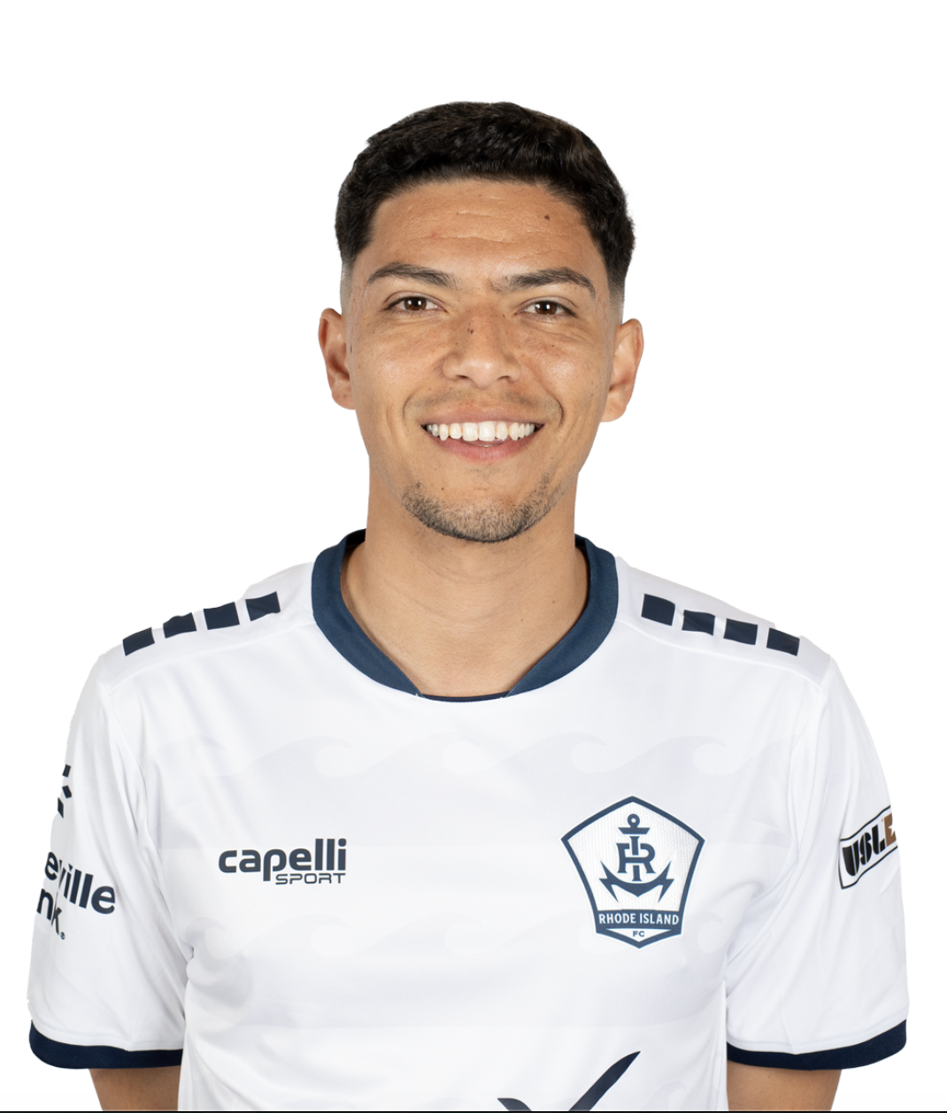 A man wearing a white Rhode Island soccer jersey, smiling, with short dark hair.