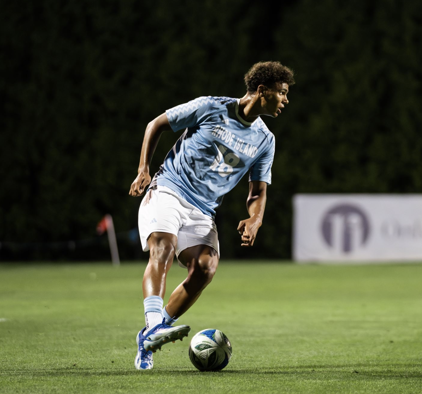 A soccer player wearing a Rhode Island team jersey and white shorts is on the field, dribbling a soccer ball during a game at night.