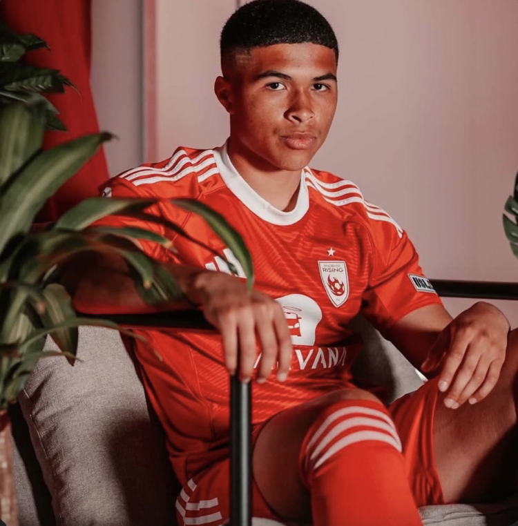 A young man wearing a red soccer jersey and shorts, sitting on a gray sofa with plants partially visible in the foreground.