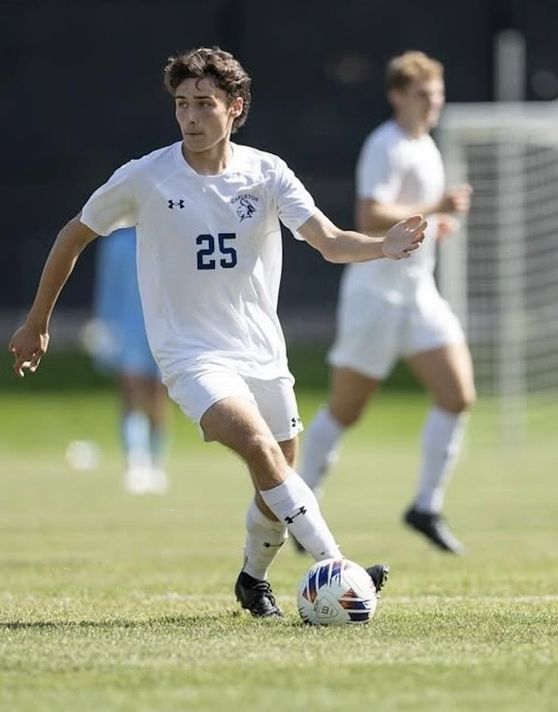 A young man playing soccer on a field, wearing a white uniform with the number 25, kicking a soccer ball. Another player is visible in the background.