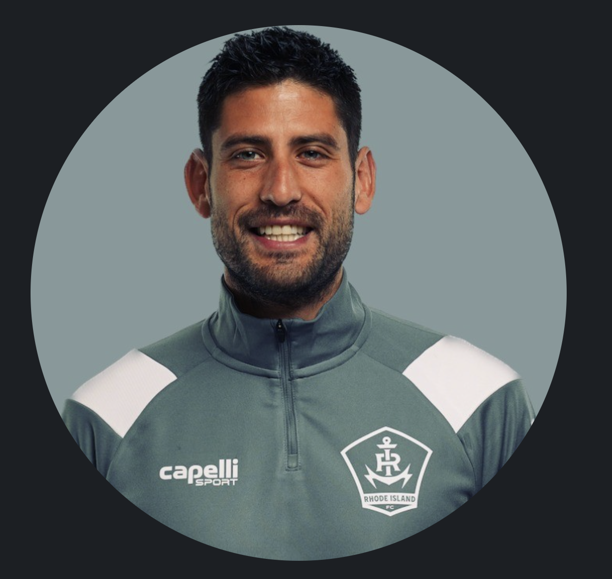 Portrait of a man with dark hair and beard wearing a gray sports jacket with a Rhode Island FC logo, smiling at the camera.