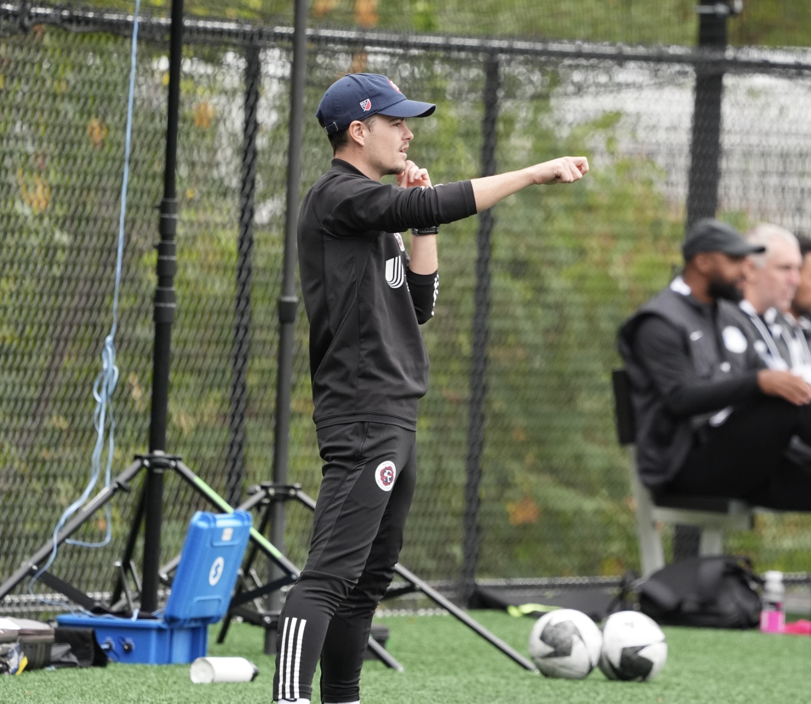 A woman with short blonde hair, wearing a sleeveless orange top and beige pants, sits on a light-colored bench and writes in a notebook with a pen. Behind her, there's a large green plant, and a framed abstract artwork is leaning against the wall. Sunlight streams through a window, casting shadows of the plant's leaves onto the wall.