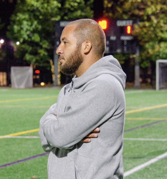 A man in a gray hoodie standing on a sports field at night, with a scoreboard in the background.
