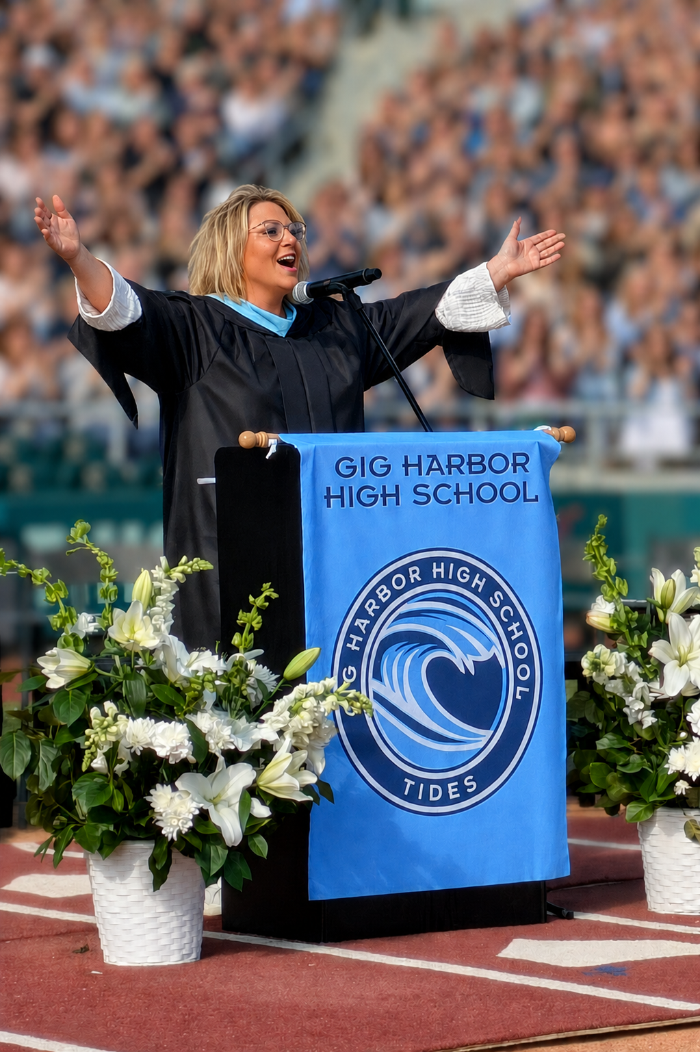 Kennedy Floyd in graduation attire standing at a podium with a blue banner that reads 'Gig Harbor High School' and a school emblem. She has her arms raised and appears to be giving a speech during a graduation ceremony.