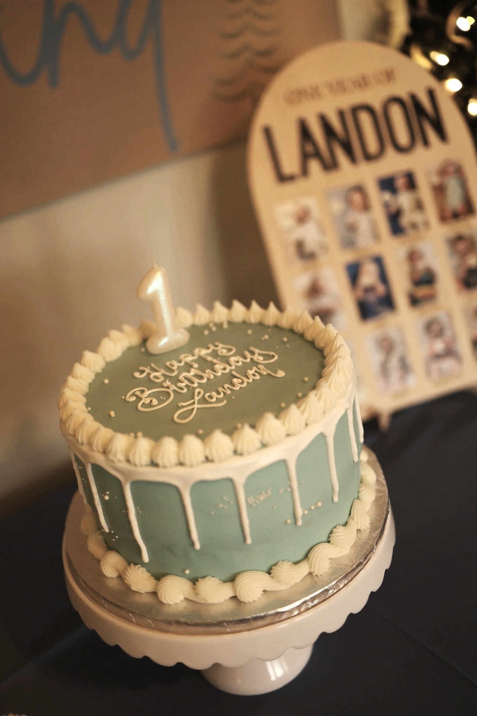 A small birthday cake decorated with light blue and white frosting, with a single candle shaped like the number one on top, and the words "Happy Birthday Landon" written on it. In the background, there is a display with photos labeled "Landon."