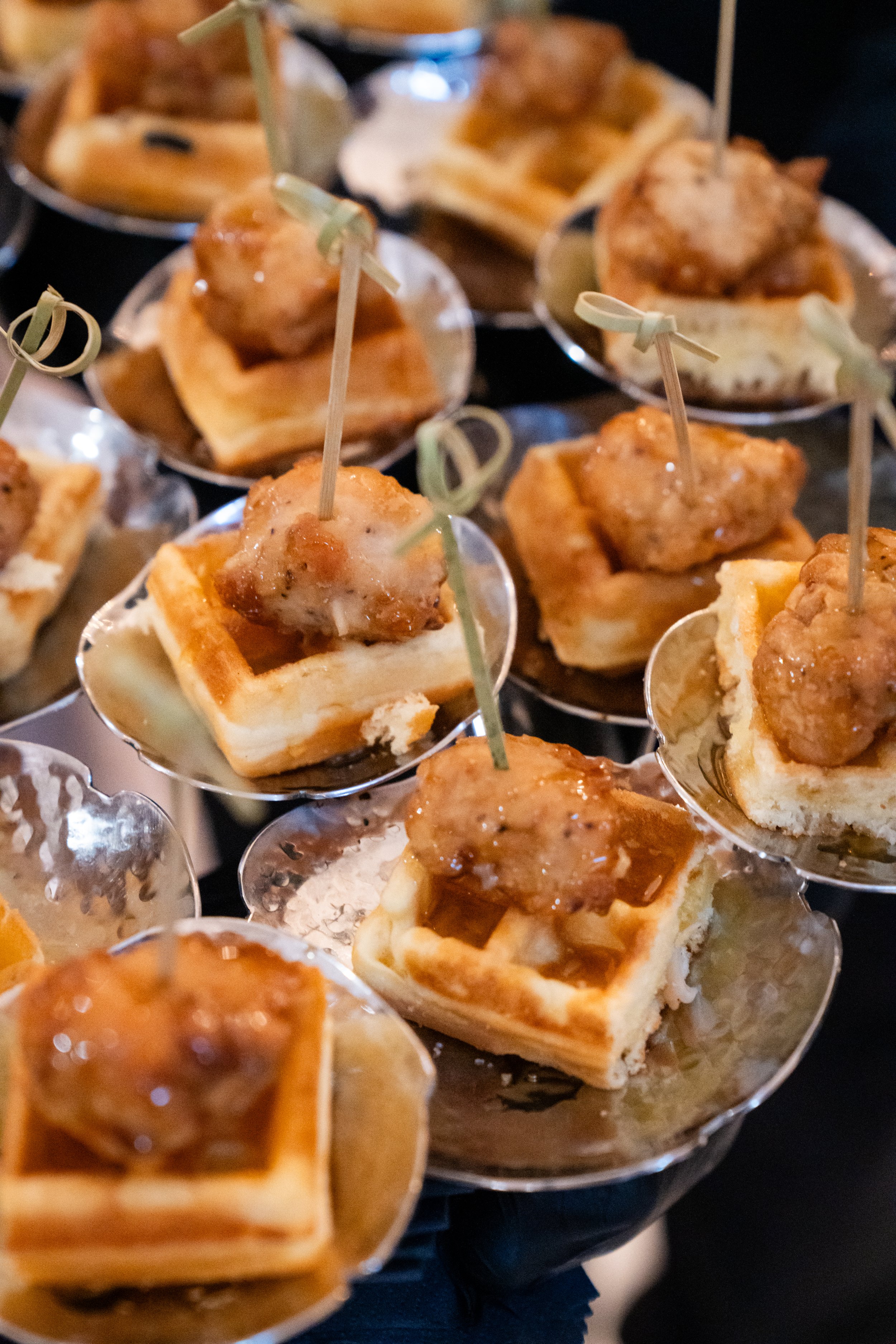 Close-up of small savory bites featuring waffle squares topped with glazed chicken pieces, served on small metallic plates with skewers.