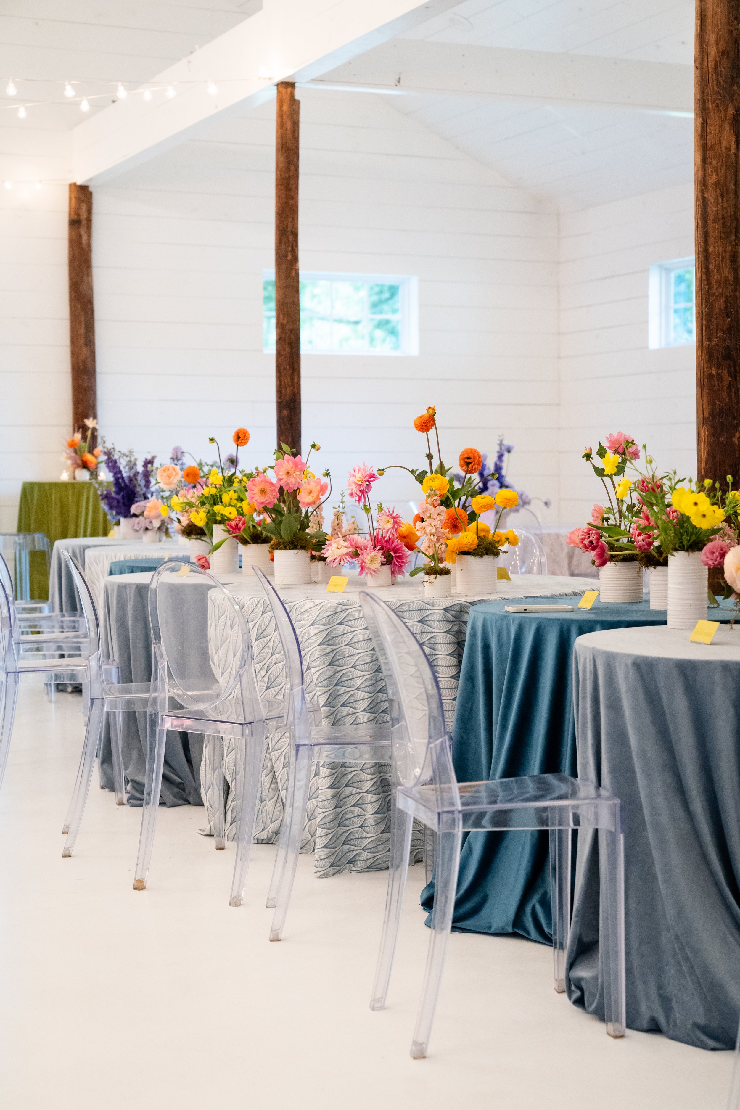 Table decorated with colorful flowers in vases, set in a bright, white room with wooden beams and clear chairs.