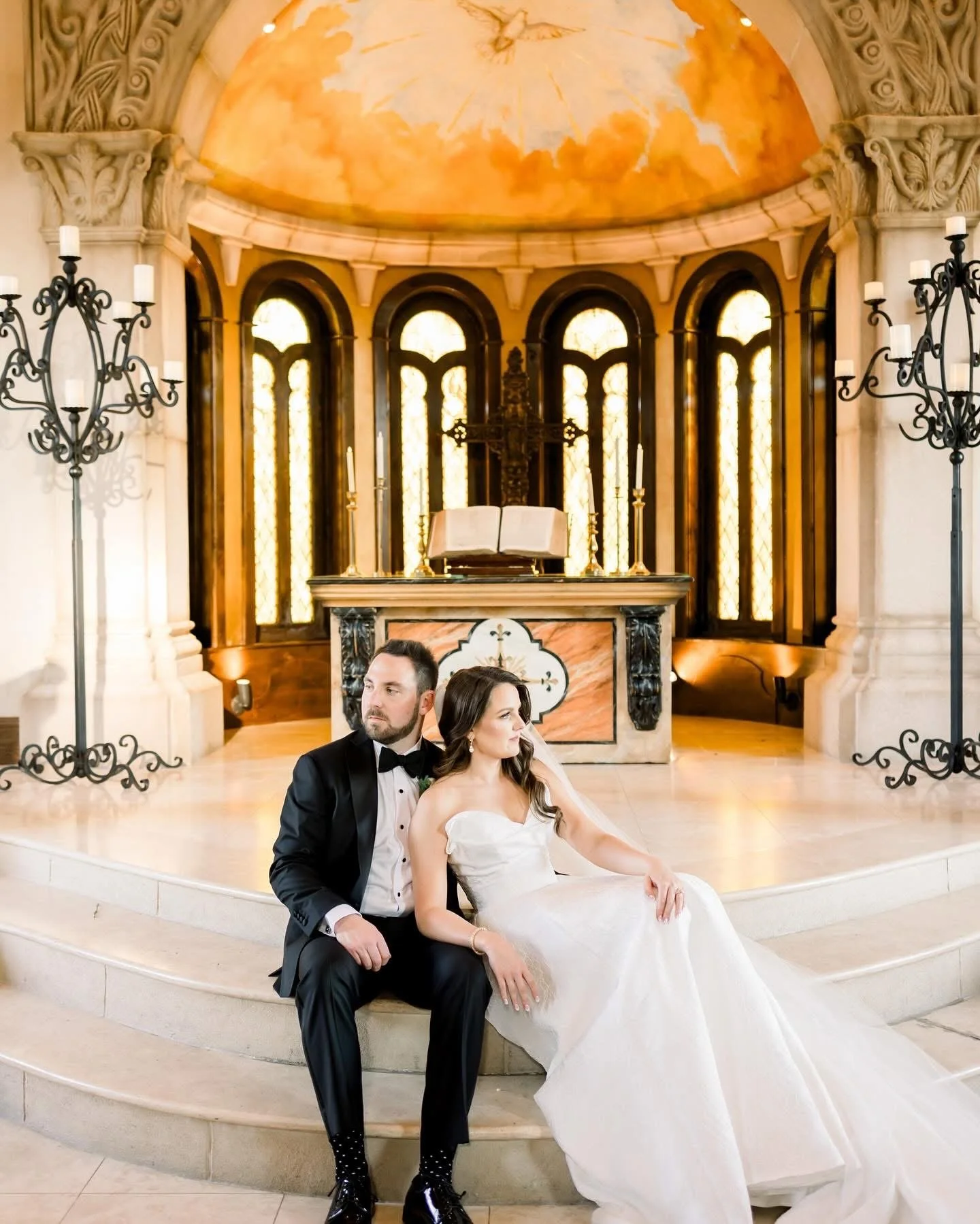 Bride and groom sitting near the altar of a church with stained glass windows and religious artifacts, dressed in formal wedding attire.