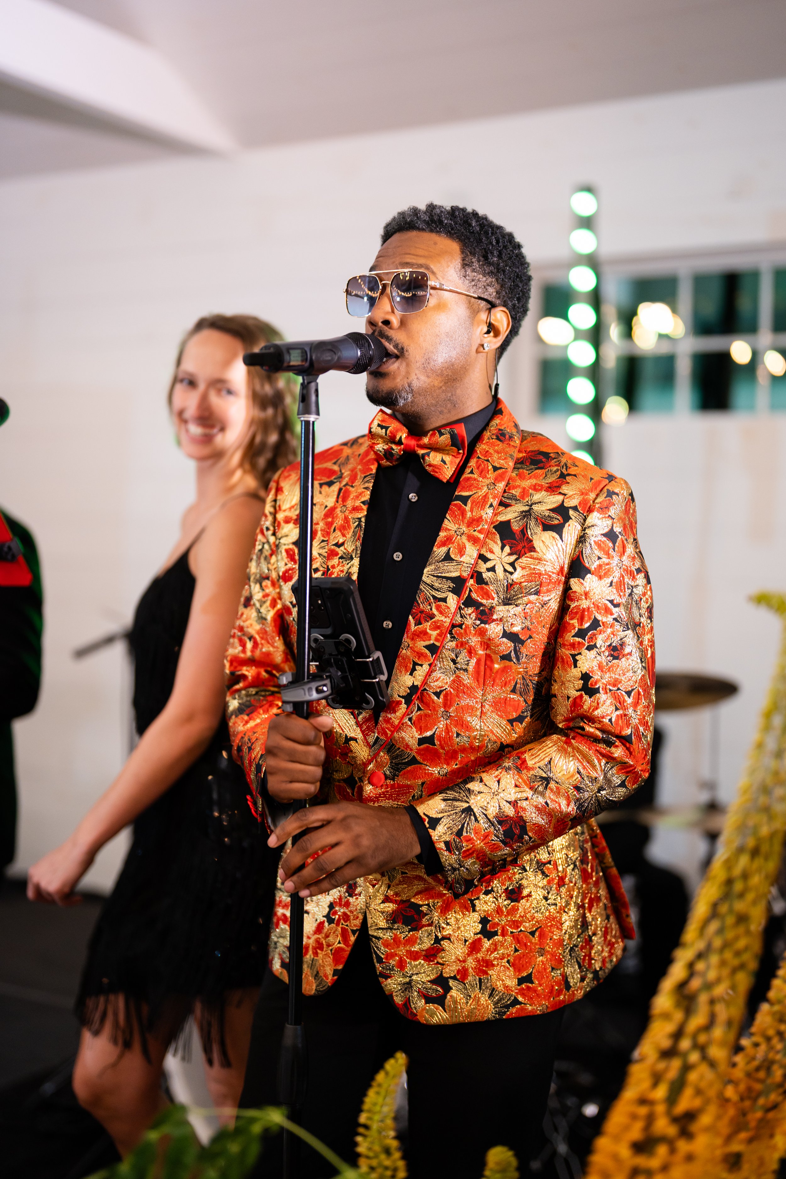 A man wearing a floral-patterned jacket and bow tie, sunglasses, and holding a microphone, appears to be singing or speaking at a festive event. A woman in a black dress is smiling in the background.