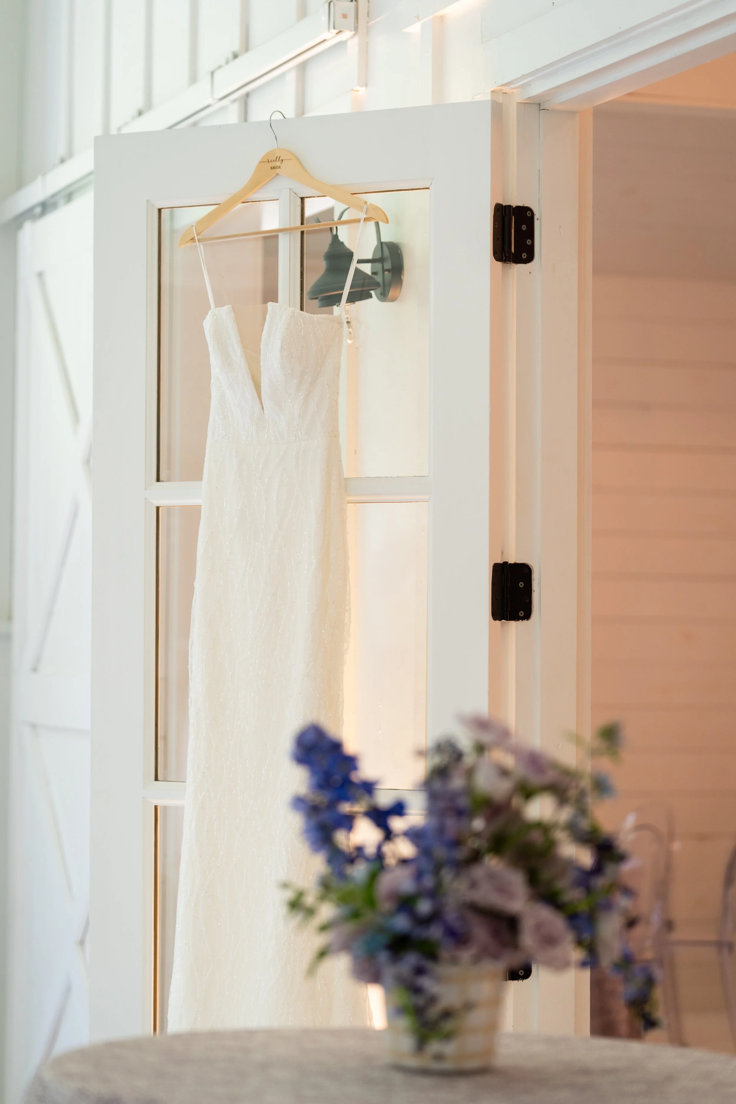 A wedding dress hanging on a hanger on a door, with a blurred flower arrangement in the foreground.