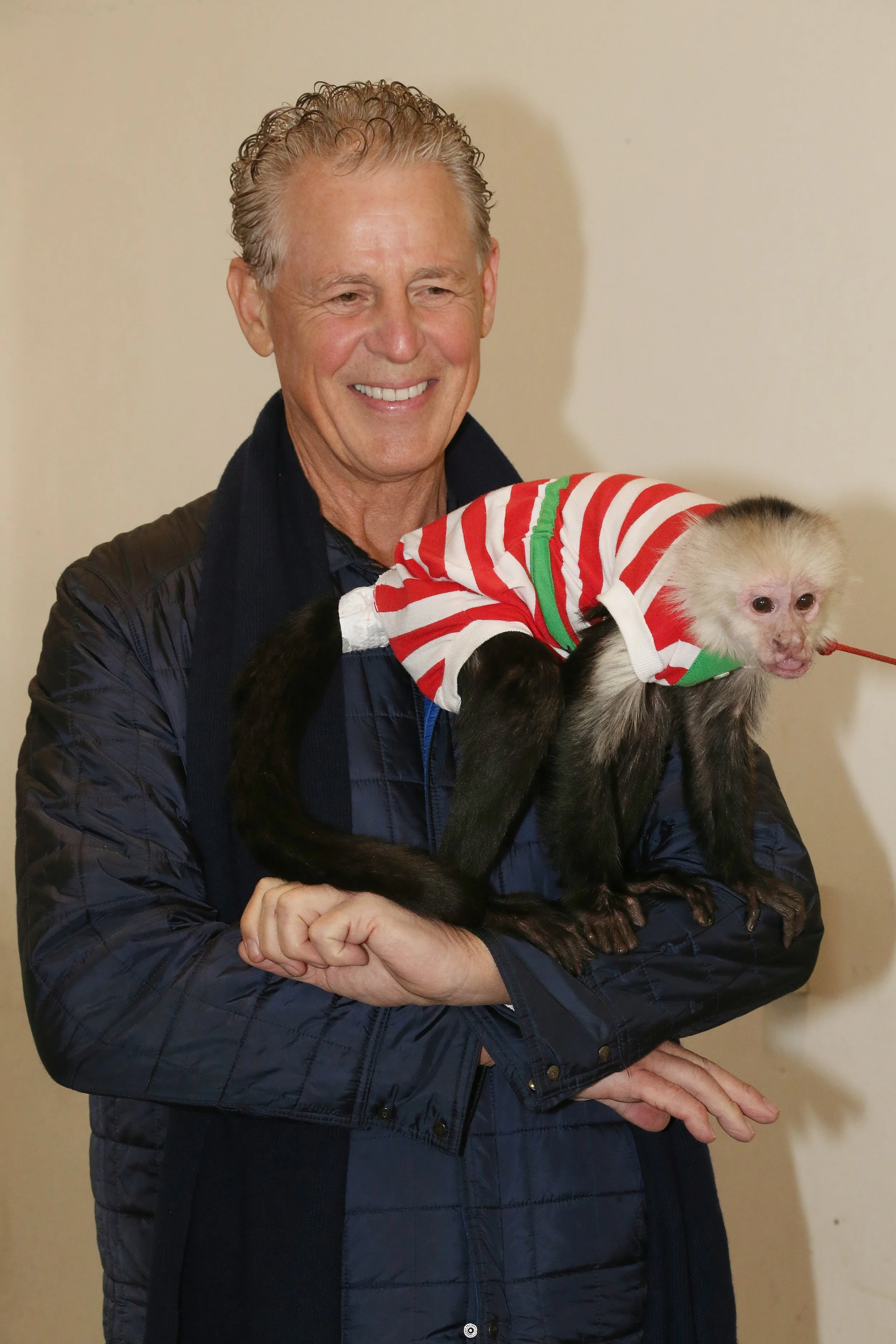 A man with gray hair and a navy jacket holds a baby monkey dressed in a white and red striped shirt with green accents.