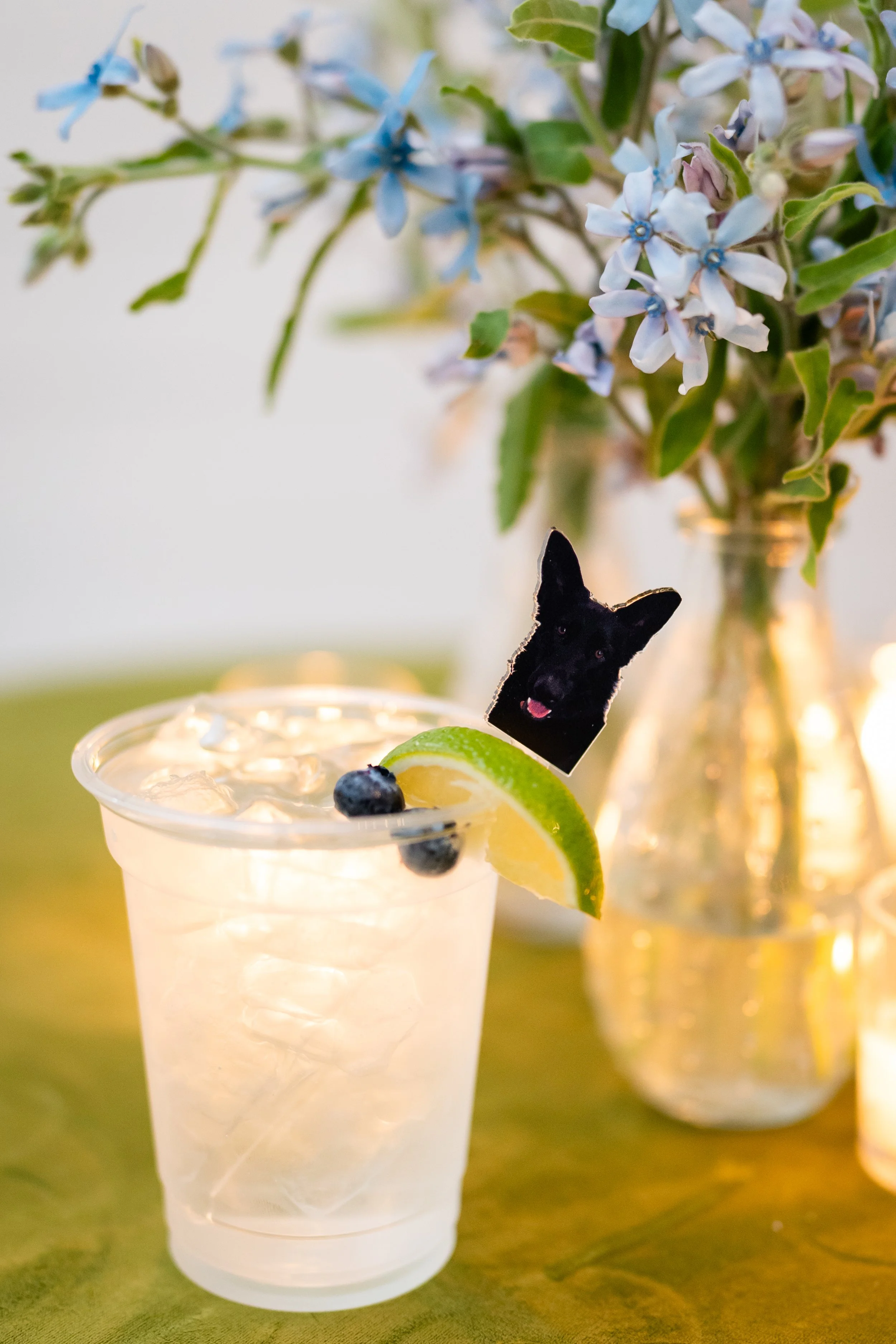 A clear plastic cup filled with a light-colored beverage with ice, garnished with a lime wedge and blueberries, and a black dog-shaped stick or decoration inside. In the background, there is a glass vase with blue flowers and green leaves.