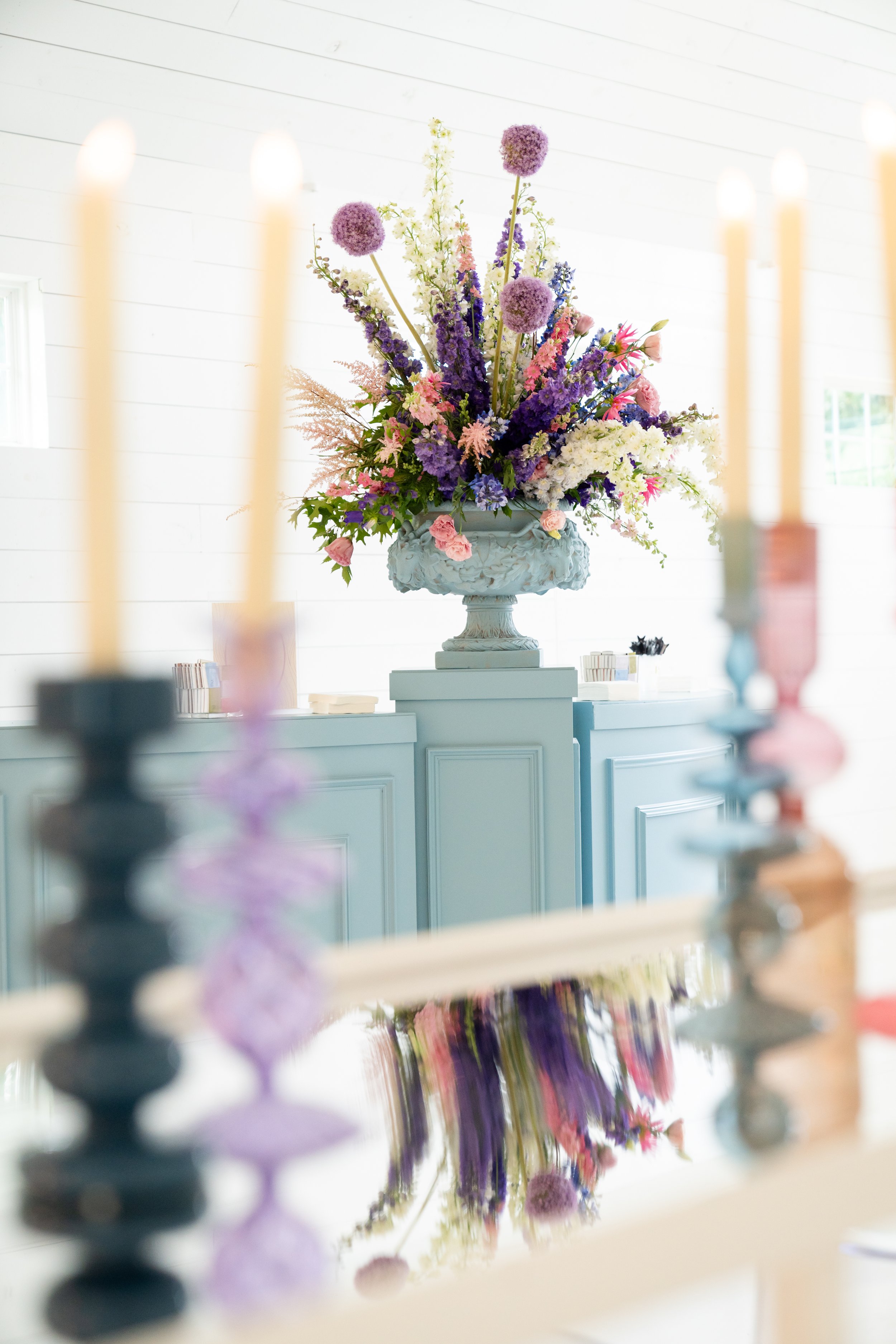 A large, ornate flower arrangement in a white vase on top of a light blue cabinet, with candles in the foreground.