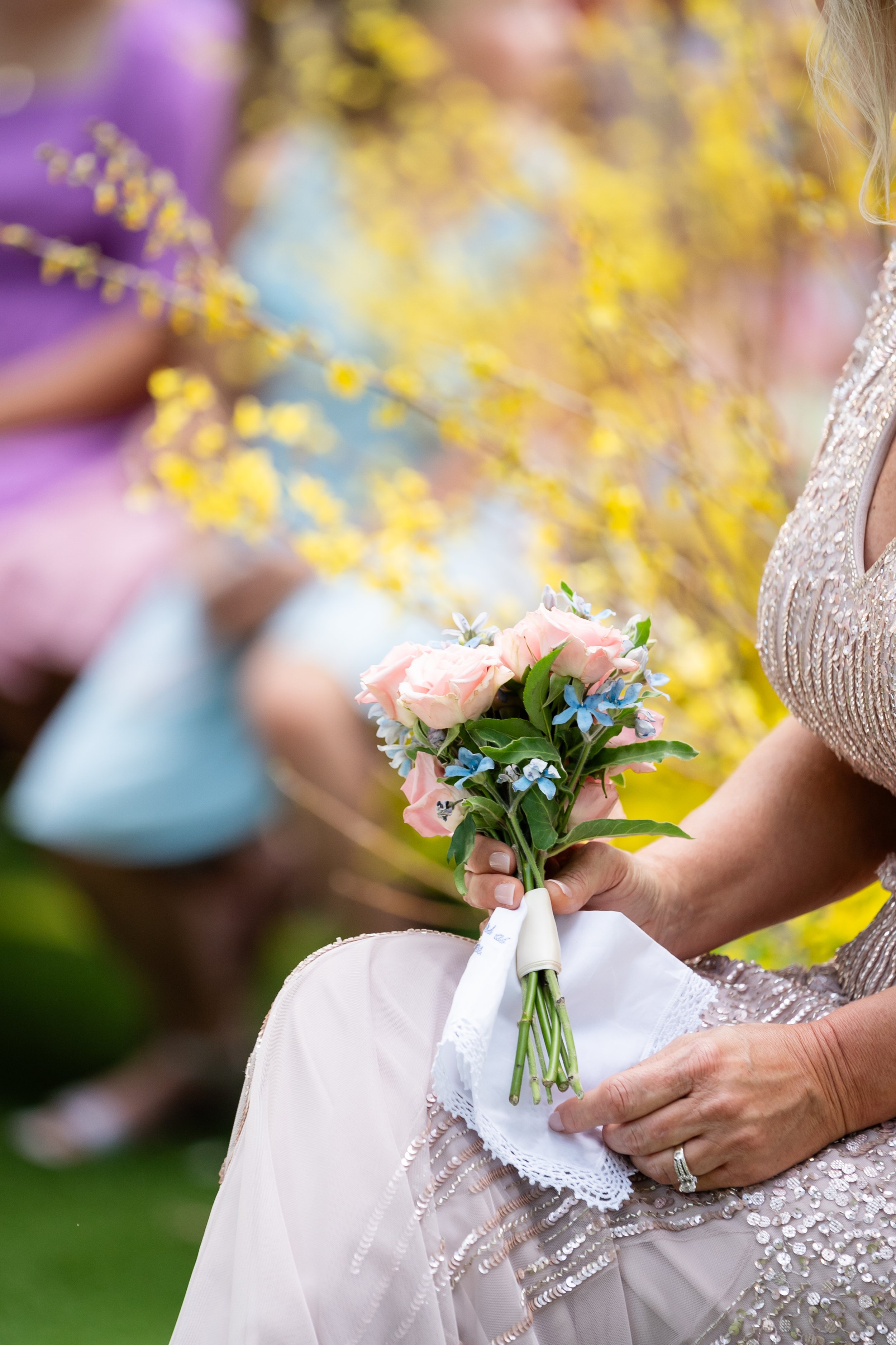 A woman in a beaded dress sitting outdoors on green grass holds a small bouquet of pink roses and blue flowers, with yellow flowers in the background.