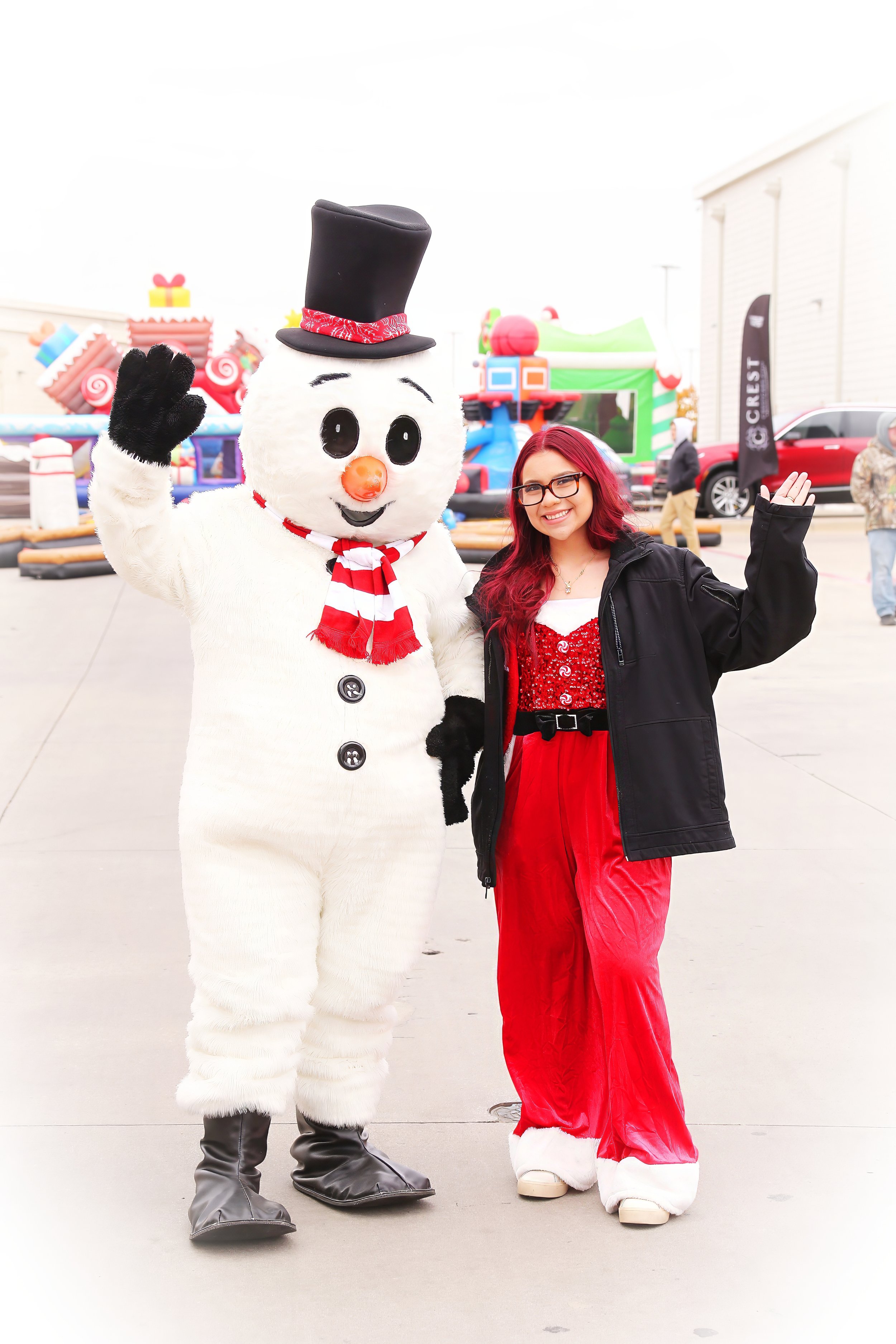 A woman in Christmas-themed red velvet pants and a black jacket standing next to a snowman mascot at an outdoor event, with inflatable holiday decorations and people in the background.