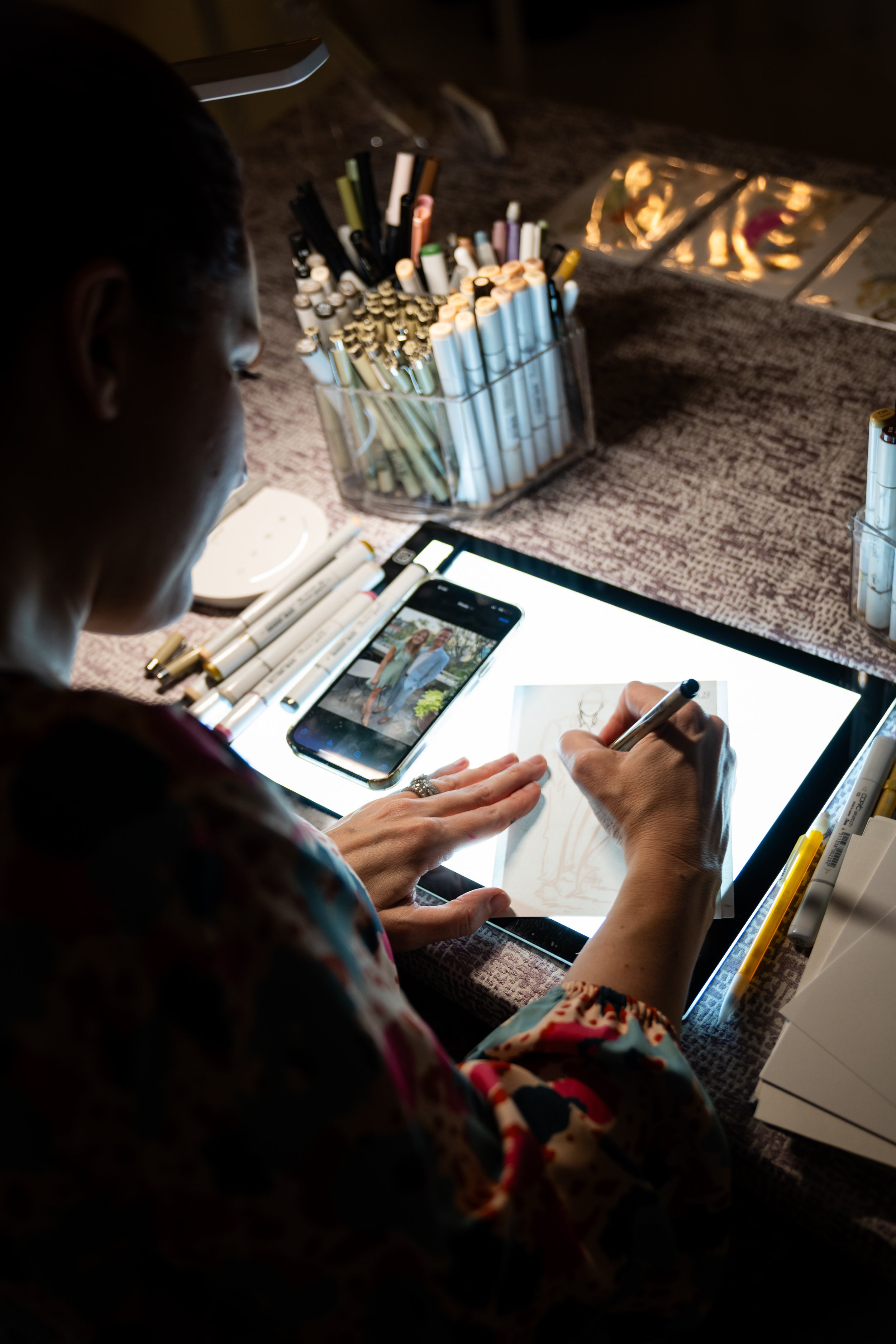 A woman seated at a table, drawing on a lightbox with markers, with a smartphone showing a photo of her and another person nearby. The table has various markers, pens, and art supplies, with additional art prints or sketches laid out on the table.