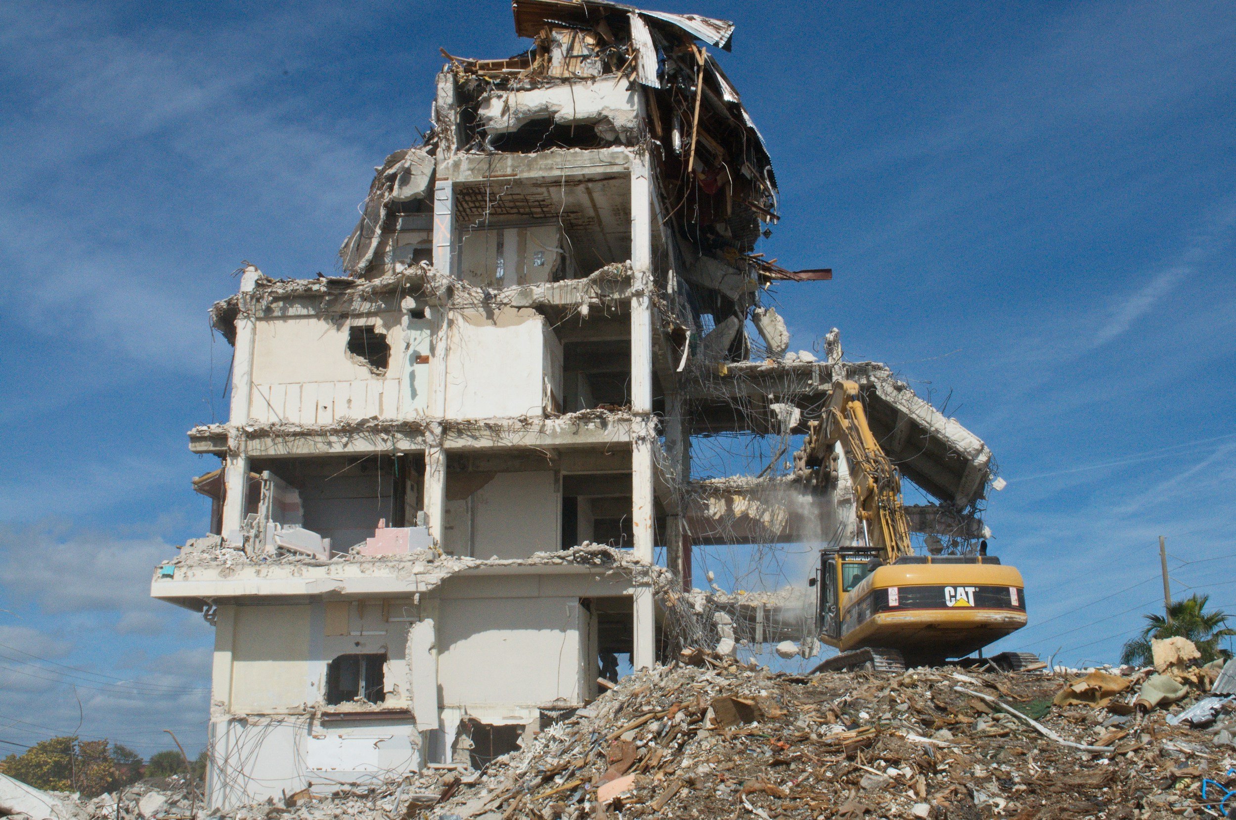 A partially demolished multi-story building with a yellow excavator on the rubble in front of it, under a blue sky.