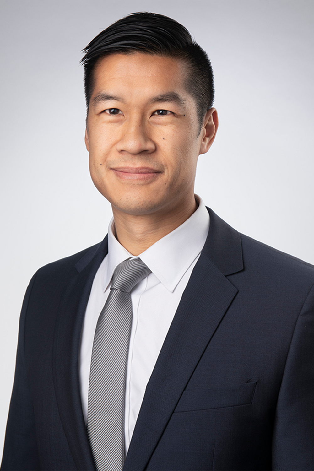 Professional headshot of a man wearing a dark suit, white shirt, and gray tie, smiling slightly, with short black hair and a light background.