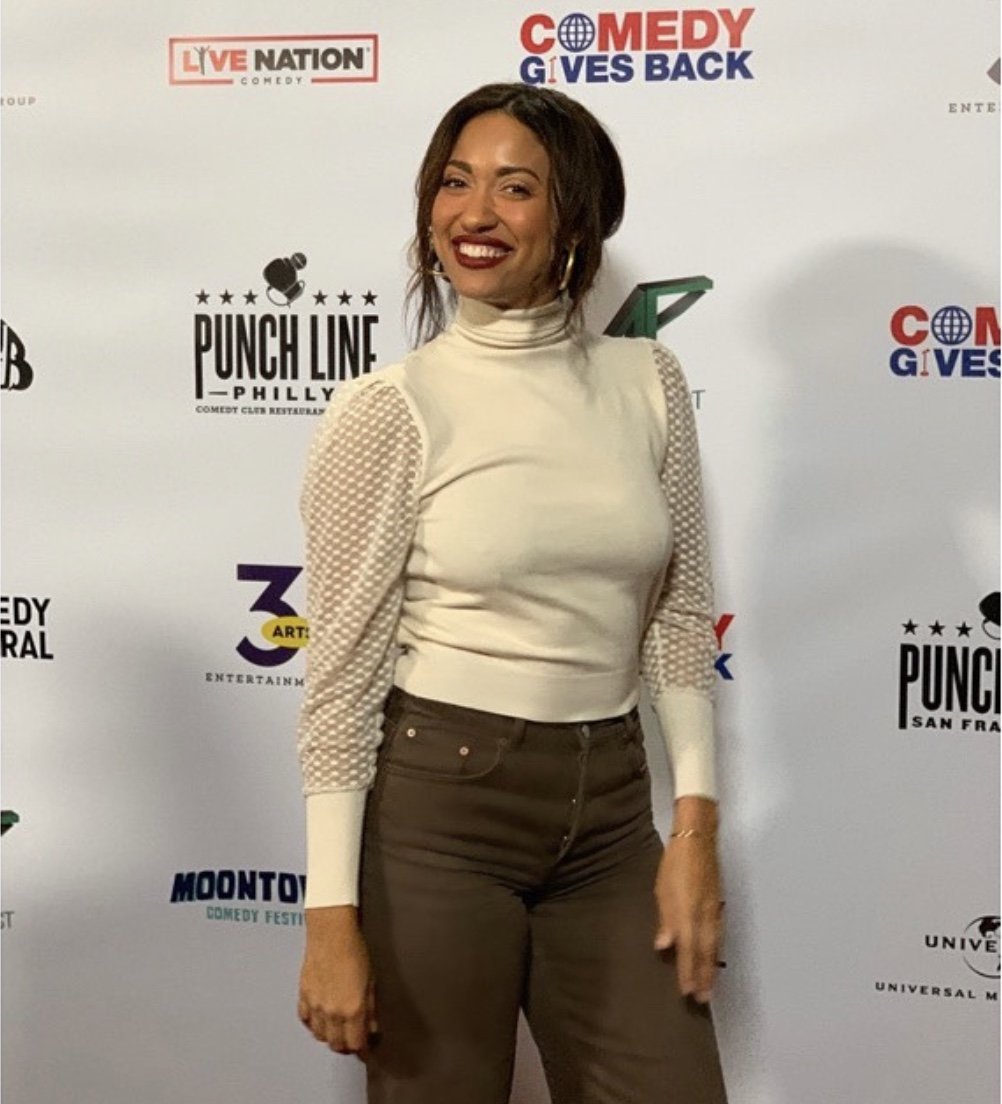 A woman smiling at a comedy event with a white background featuring multiple logos, including 'LIVE NATION,' 'COMEDY GIVES BACK,' and 'PUNCH LINE PHILLY.'