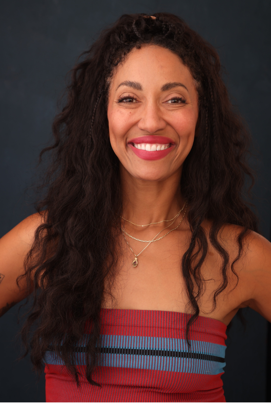 Smiling woman with curly dark hair wearing a strapless red top with blue and black stripes, layered necklaces, and a small gold bracelet.