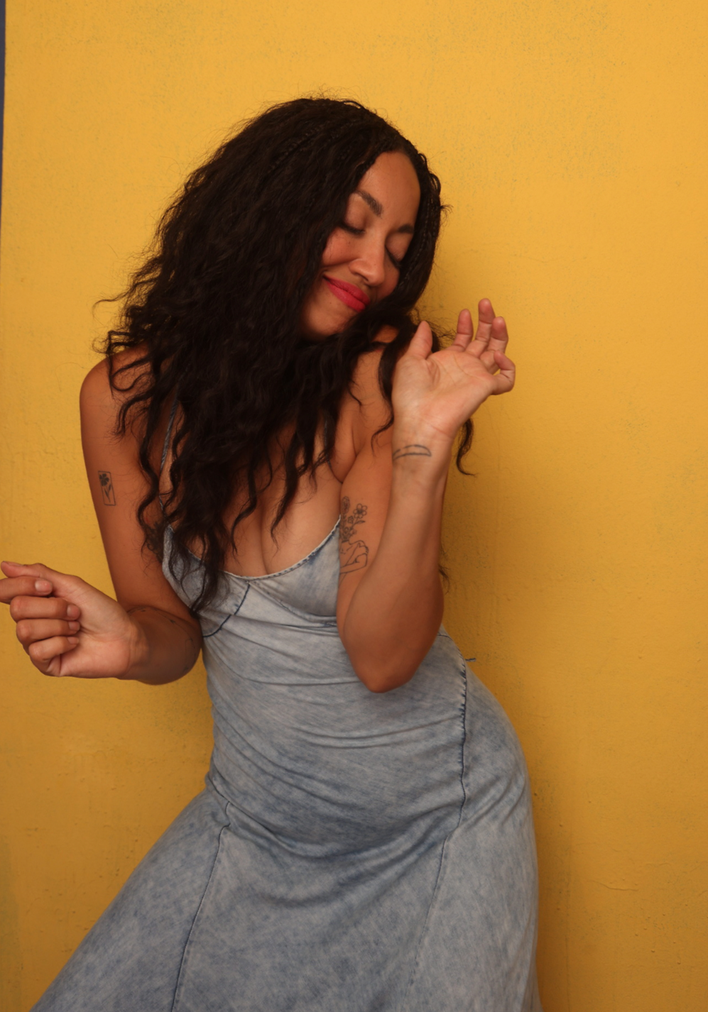 A woman with long curly hair, tattoos on her arms, wearing a grey denim dress, is dancing with a smile against a yellow wall.
