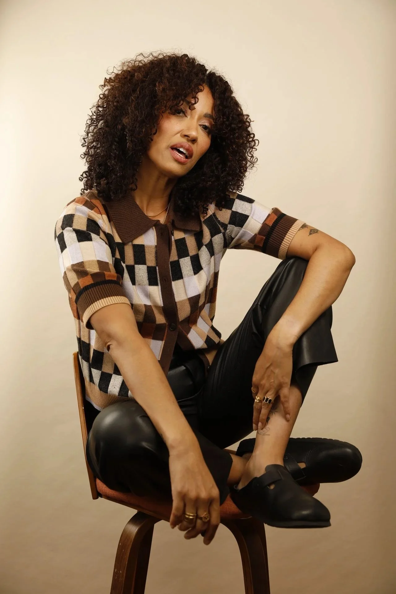 A woman with curly hair sitting on a chair, wearing a checkered shirt, black pants, and black shoes, against a plain beige background.