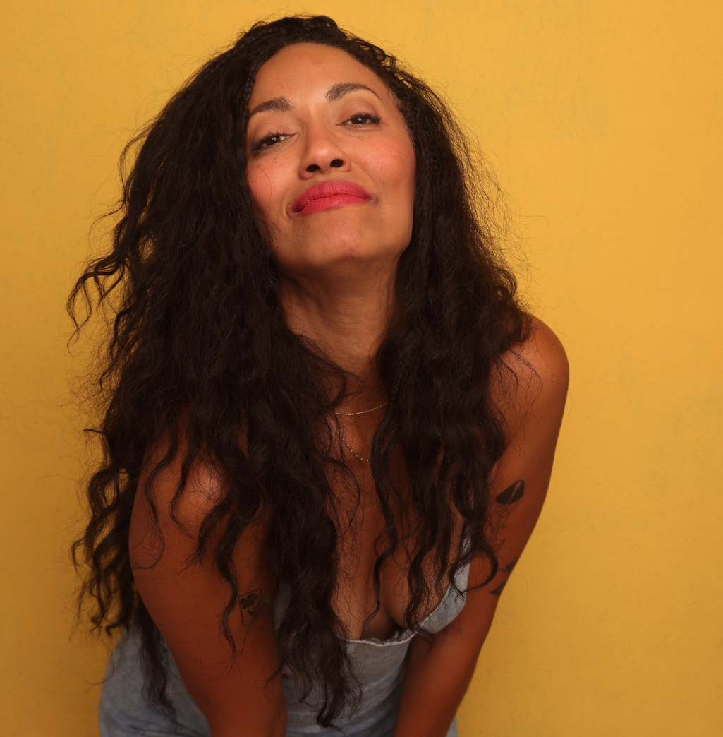 A woman with long, curly dark hair and red lipstick leans forward slightly, looking directly at the camera, against a yellow background.
