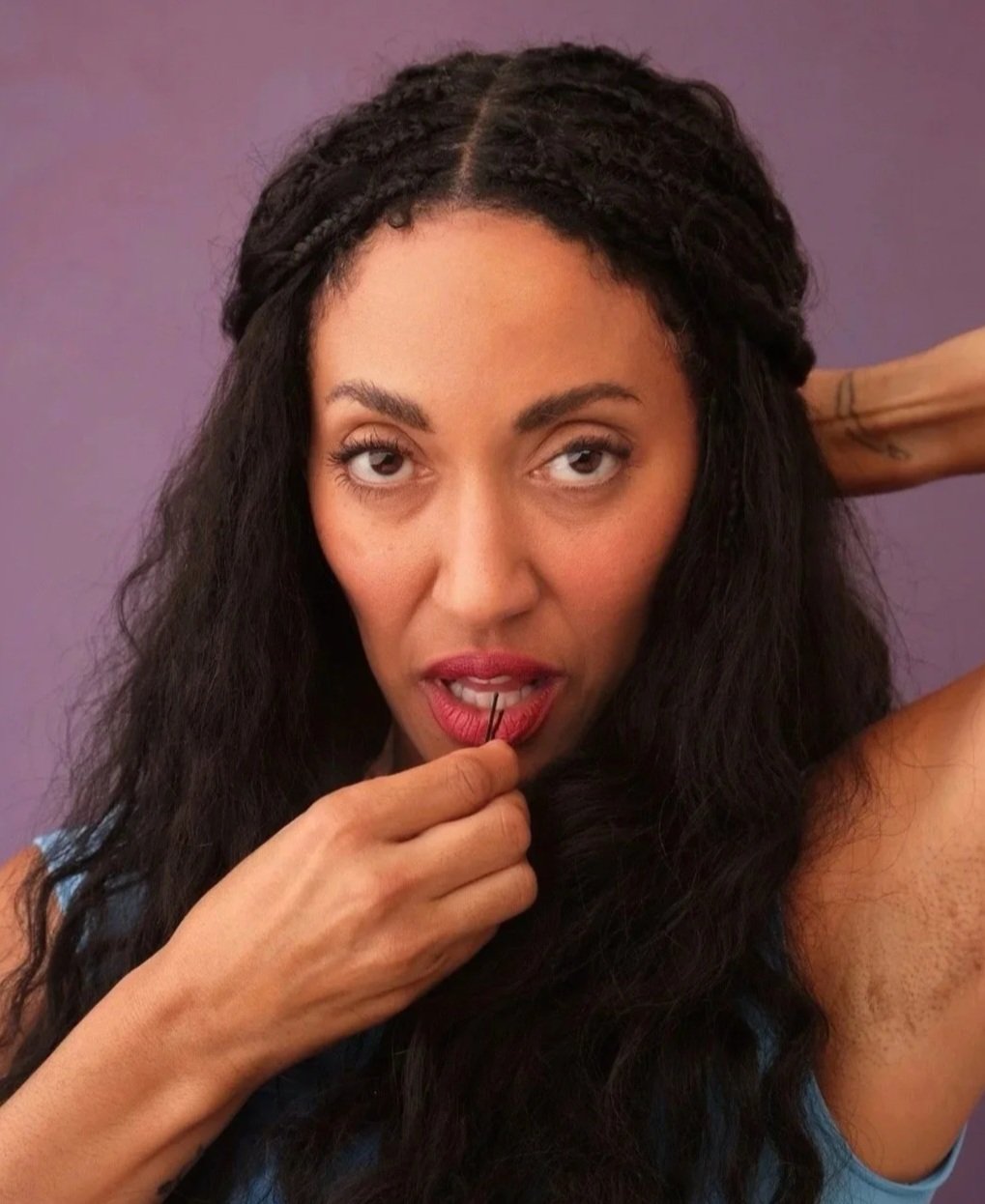 A woman with long curly black hair, holding a hairpin near her lips, looking directly at the camera with a slightly serious expression, and a purple background.