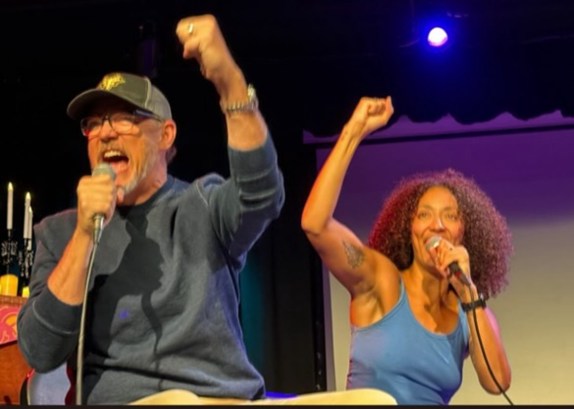 Two people, a man and a woman, are on stage holding microphones and raising their fists in the air, appearing joyful and energetic, performing together with a dark background and stage lighting.