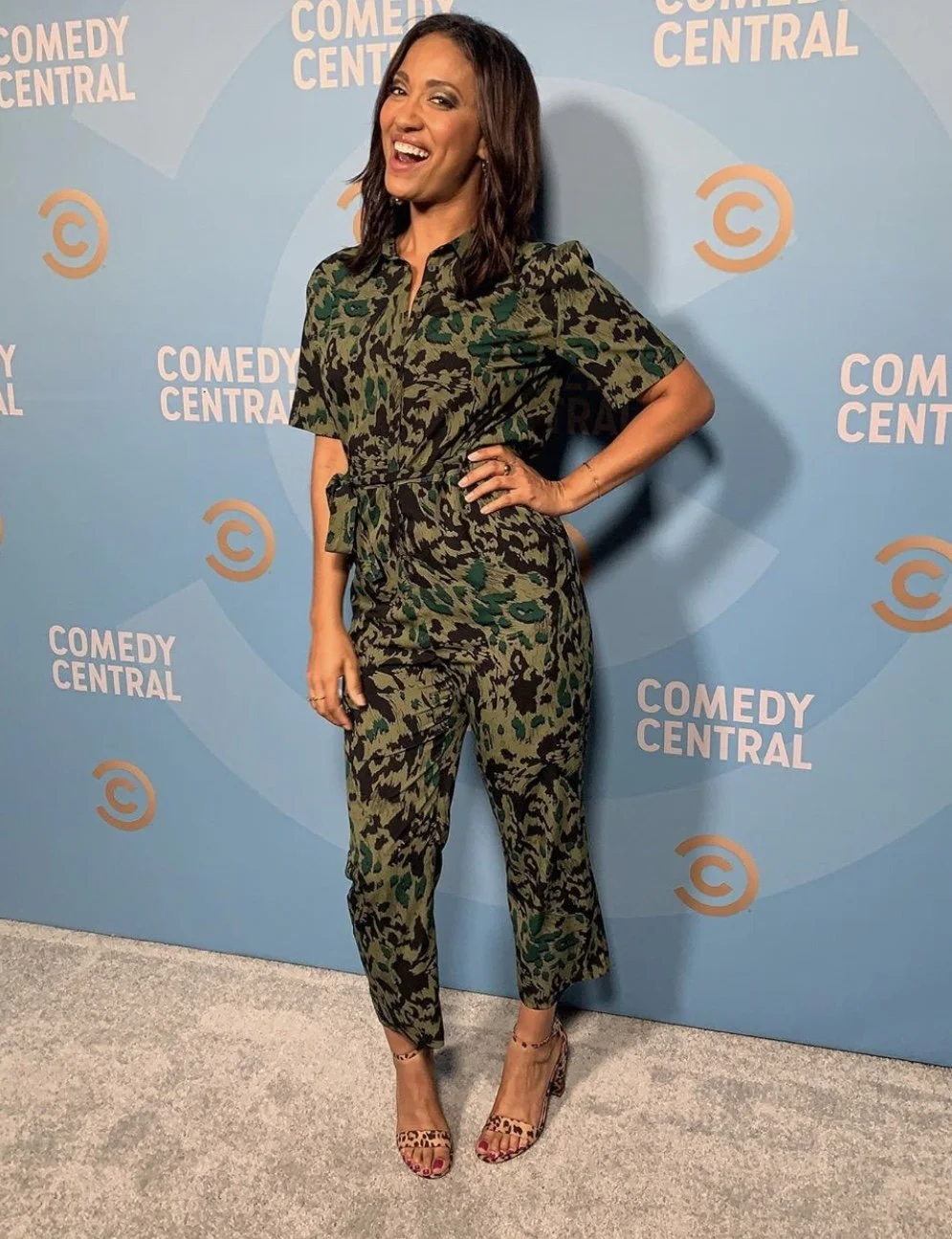 A woman standing in front of a light blue Comic Central backdrop, smiling and posing with one hand on her hip. She is wearing a matching camo print jumpsuit and leopard print high heels.