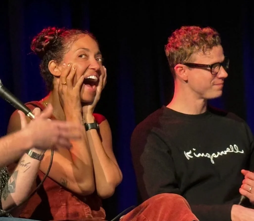 A woman with curly hair in buns expressing surprise or excitement, sitting next to a man with glasses, wearing a black shirt with gold lettering, on a stage with a blue curtain background.