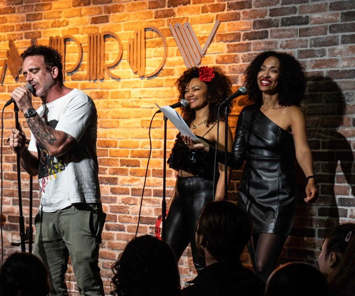 A man singing into a microphone on stage with two women, one holding a script, in front of a brick wall with the word 'STANDREW'S' visible behind them. The women are smiling and wearing black outfits, and the audience is visible in the foreground.
