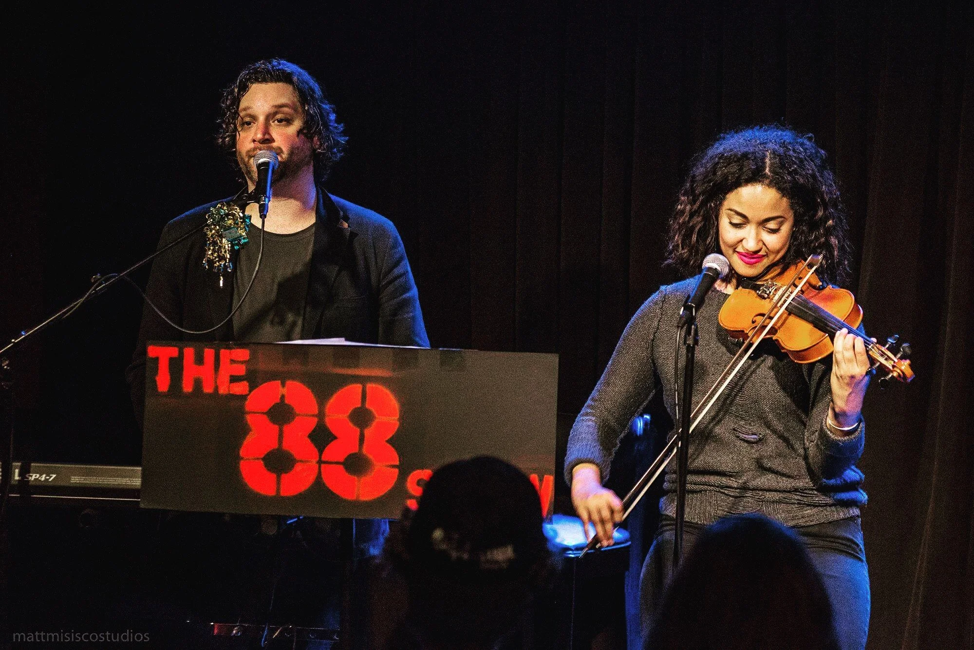 A male musician with curly hair and a black outfit stands at a keyboard with a microphone, and a female violinist with curly hair and a dark sweater plays the violin, both performing on stage at The 88 show.