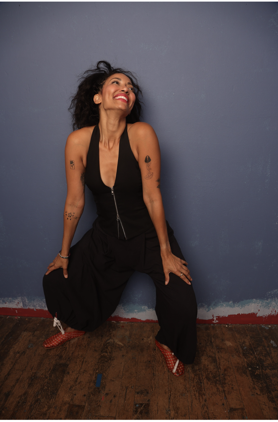 A woman with curly black hair, wearing a black sleeveless zip-up top and black loose pants, standing against a blue wall. She is smiling with her head tilted back and hands on her thighs, wearing red fishnet high heels.
